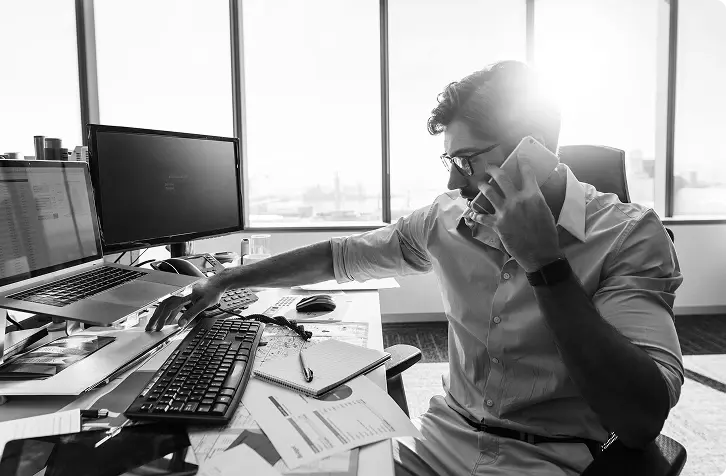 A man speaking on the phone while working at a desk with monitors and paperwork.