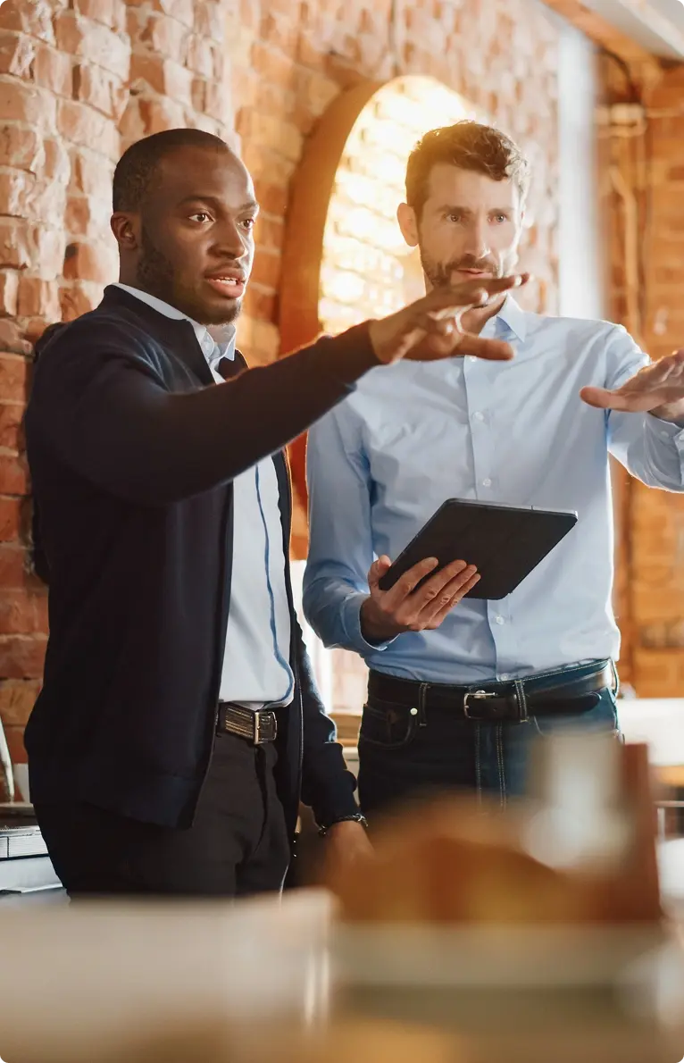 Two men talking in a brick-walled workspace, one holding a tablet.