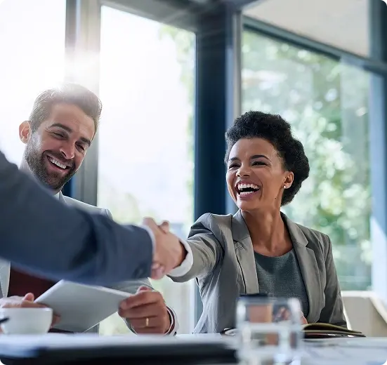 A woman shaking hands across a meeting table while a colleague looks on.