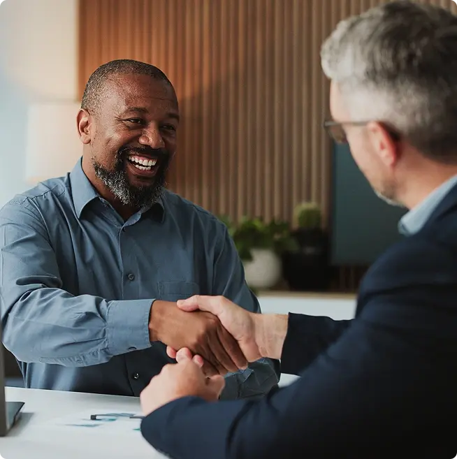 Two men shaking hands across a table during a meeting.